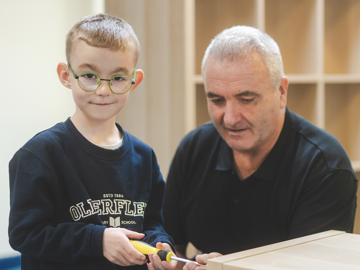 A young boy with glasses and a black shirt standing next to an older man in a black shirt. The boy is holding a yellow screwdriver and the man is assisting him. They are indoors, with wooden shelving in the background.