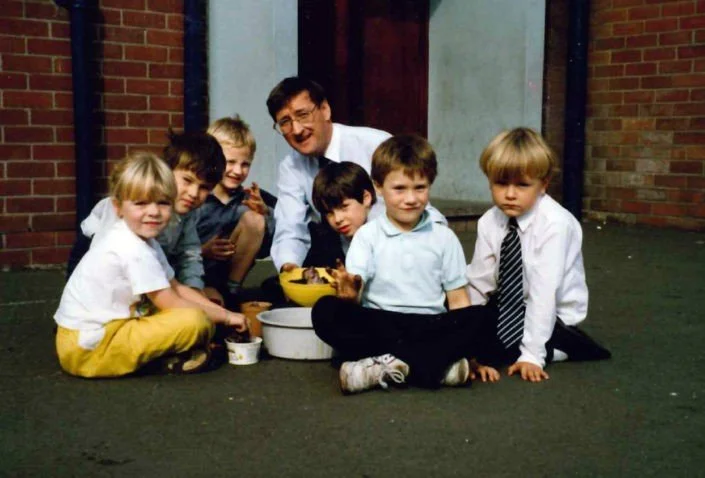 A man and six children sitting on the ground outside near a brick wall, with a yellow bowl and food, smiling at the camera.