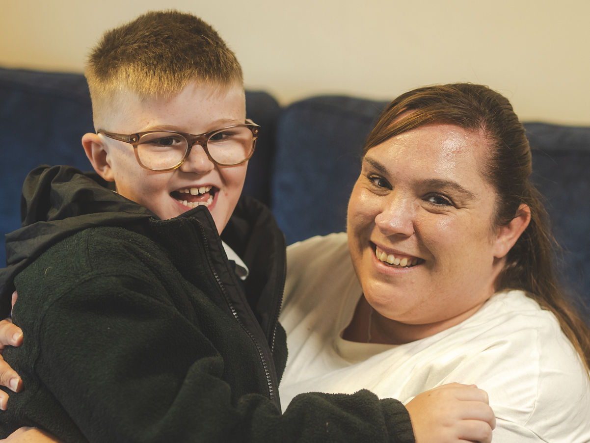A woman and a young boy smiling and embracing each other, sitting on a couch.