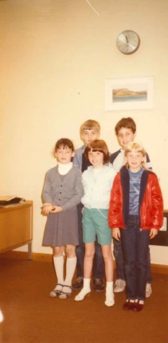 Group of five children standing in a room with a clock and framed picture on the wall.