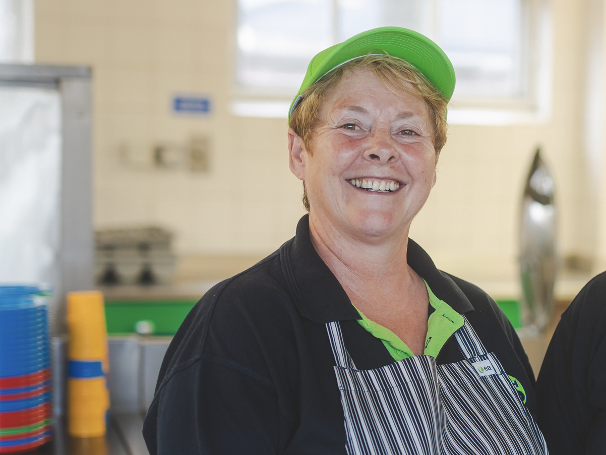 Smiling woman wearing a green cap, black polo shirt with a green collar, and striped apron in kitchen environment.