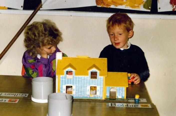 Two children playing with a wooden toy house at a table.
