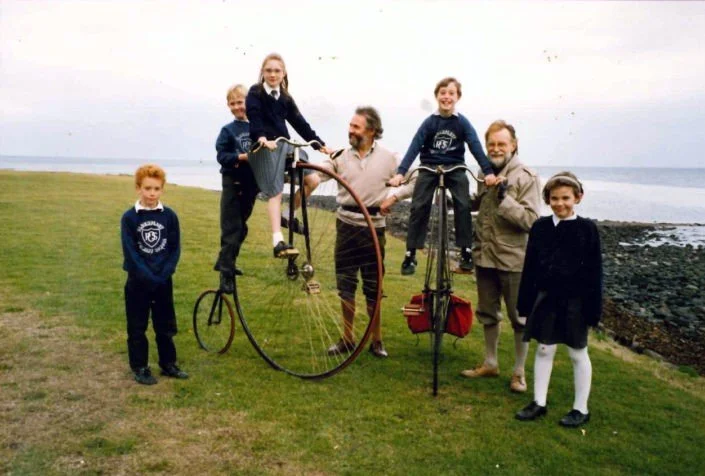 A group of five children and two adults near a beach with a grassy area and rocky shoreline, some children riding penny-farthings, and everyone smiling.