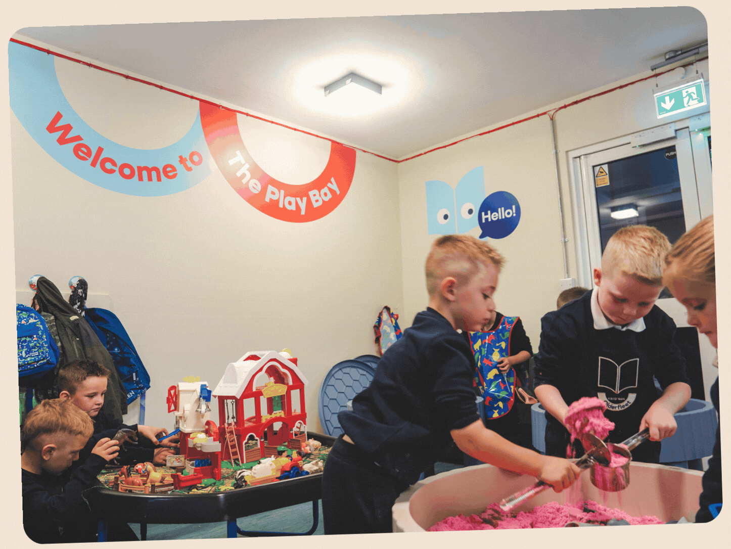 Children playing with toys and sand toys in an indoor play area with a sign reading 'Welcome to The Play Bay' and cartoon book character on the wall.
