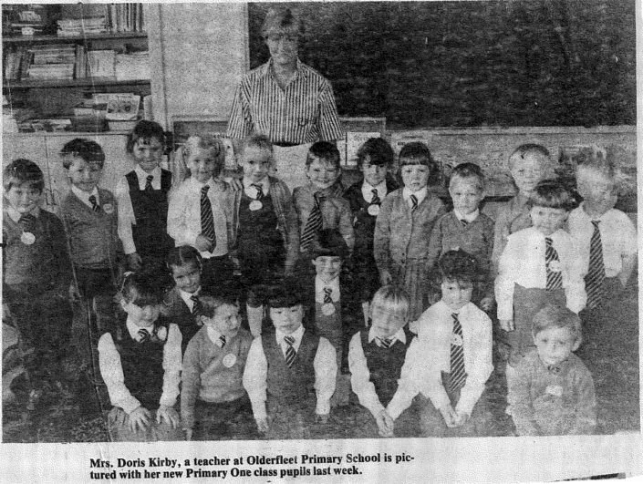Black and white photo of Mrs. Doris Kirby, a teacher at Olderfleet Primary School, standing with her new Primary One class pupils in a classroom with bookshelves and a chalkboard behind them.