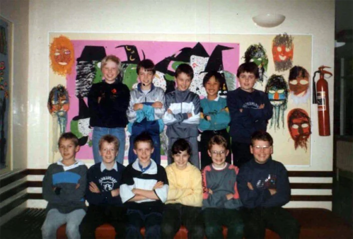 Group of ten children, five standing and five sitting, smiling in front of colorful artwork of masks on a wall, with a fire extinguisher on the right.