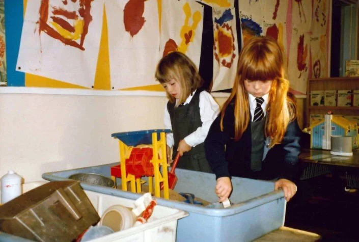Two young girls in school uniforms playing in a classroom with a toy play kitchen set, standing at a large blue basin filled with water and dishes, walls decorated with colorful artwork.
