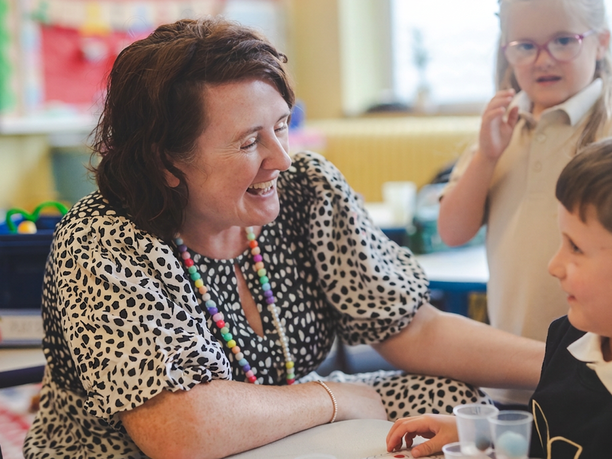 A woman with short brown hair and a colorful beaded necklace smiling and engaging with two children in a classroom setting.