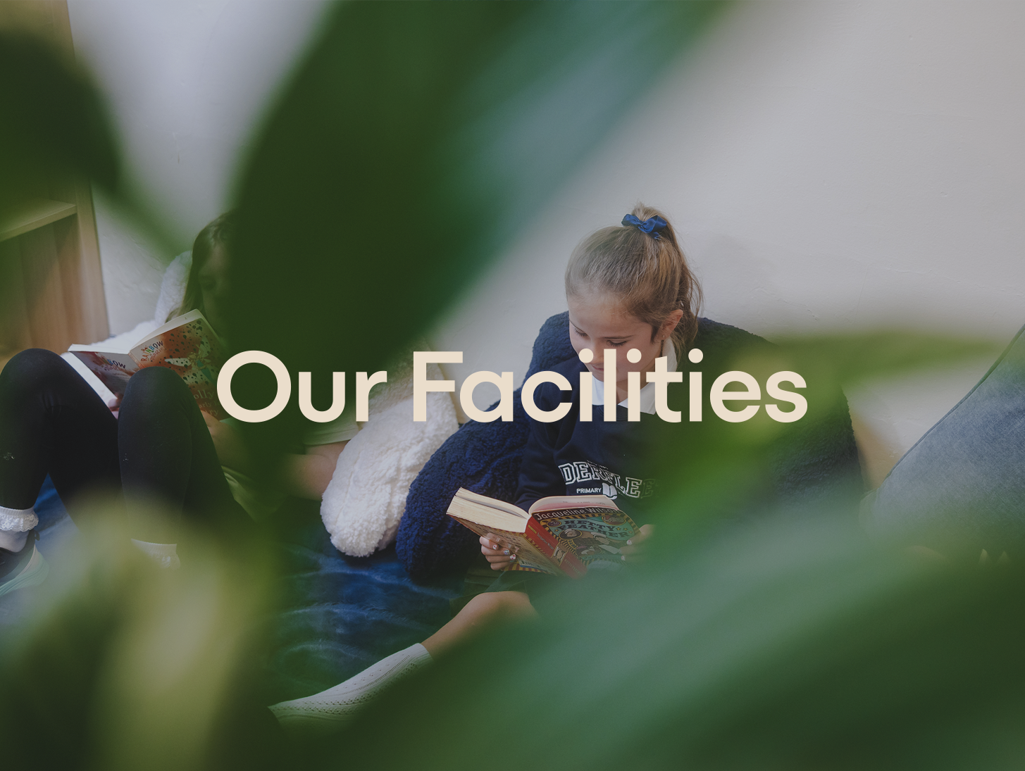 Two children sitting on a soft surface, reading books, with green leaves partially obscuring the view and the text 'Our Facilities' overlaid on the image.