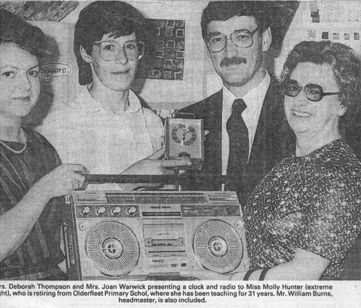 Four people, three women and one man, are standing together. One woman is holding a clock and radio. There is a large stereo with two speakers in front of them. The scene appears to be a presentation or retirement event.