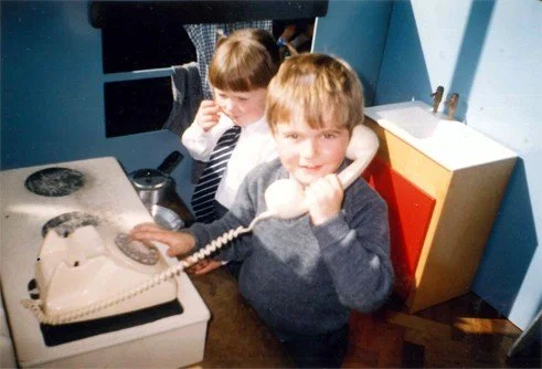 Two children, a girl and a boy, using a vintage rotary phone in a kitchen. The boy is speaking into the phone, and the girl is talking on a different phone receiver.