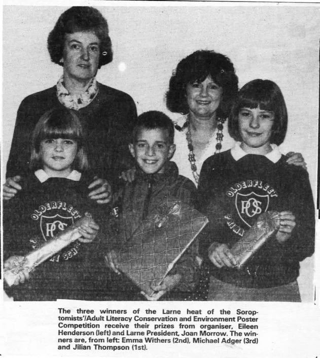 Black and white photo of three children and two women, with the children holding wrapped gifts. The children wear school uniforms with a logo that reads "Olderflich" and "PS". The women stand behind the children, smiling. Caption describes the children as winners of the Larne heat of a competition, receiving prizes from organisers.