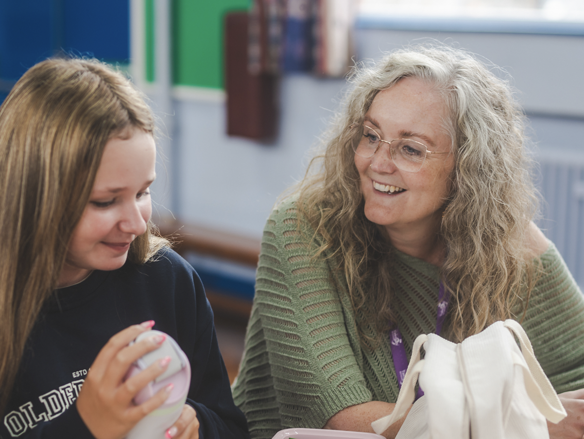 A young girl with brown hair in a dark sweatshirt smiling while holding a white object, sitting next to an older woman with curly gray hair and glasses, wearing a green knit sweater and smiling.