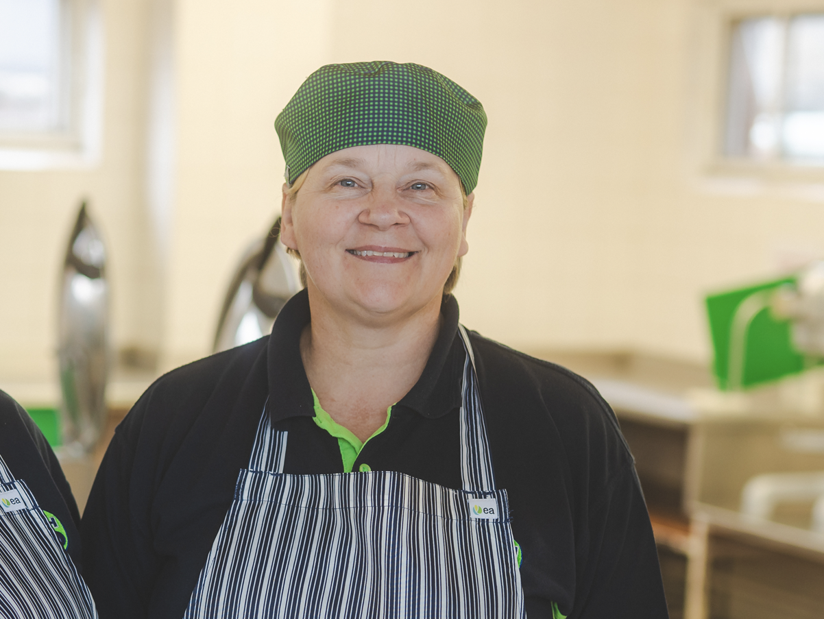 Smiling woman wearing a green and black chef hat and striped apron in a kitchen.