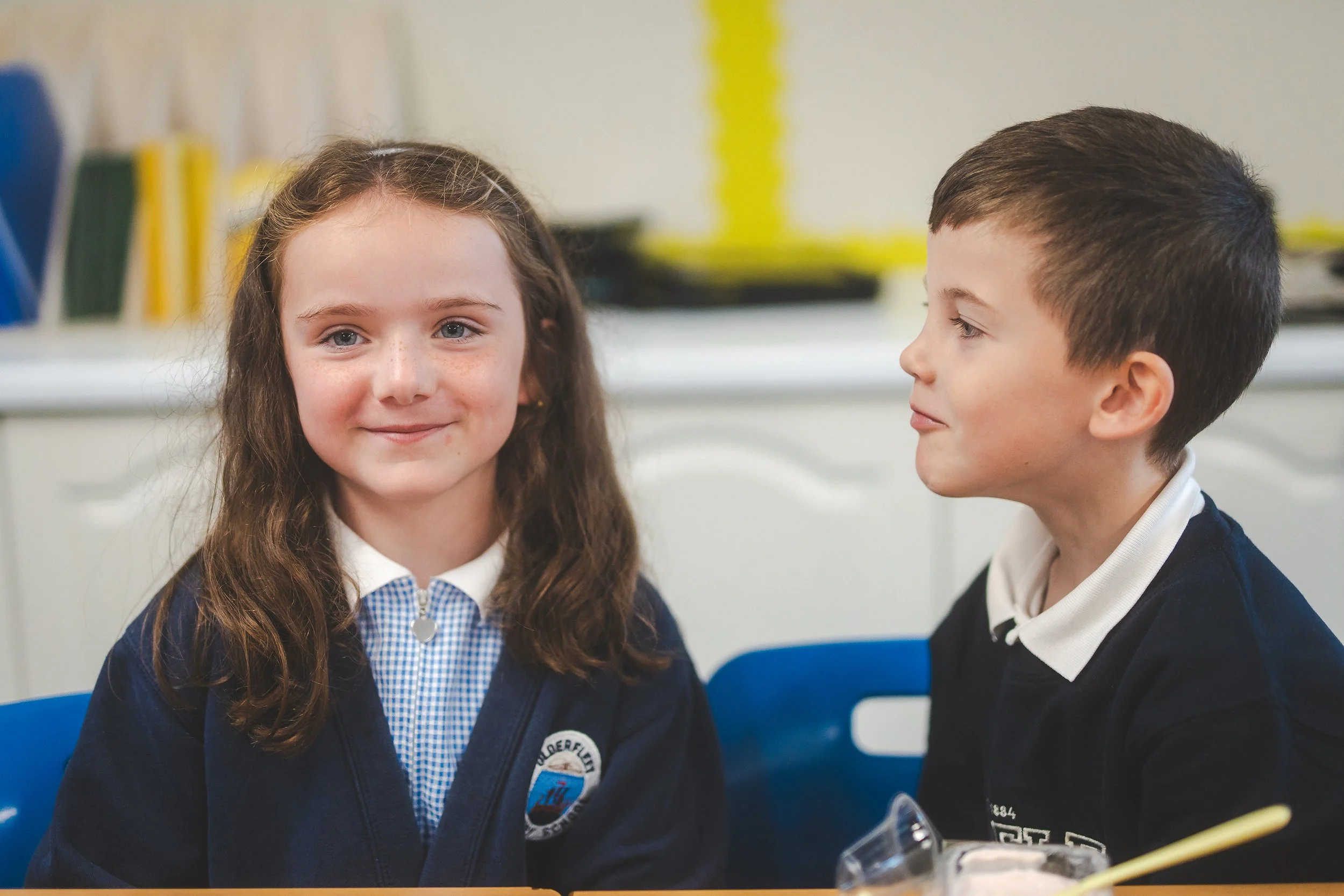 A young girl with long, curly brown hair and blue eyes smiling at the camera, wearing a school uniform and sitting next to a boy with short dark hair, also in a school uniform, in a classroom setting.