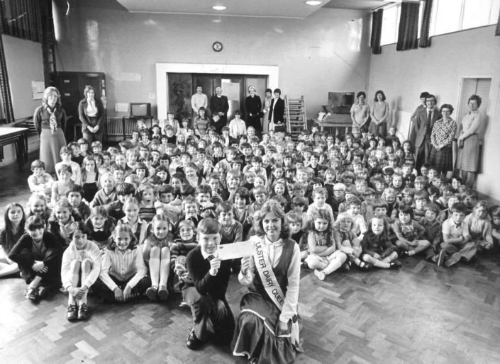 Group of children and adults in a school assembly or event, with a girl wearing a sash that says 'Student Dignity Queen' holding a certificate, in a gymnasium or hall.