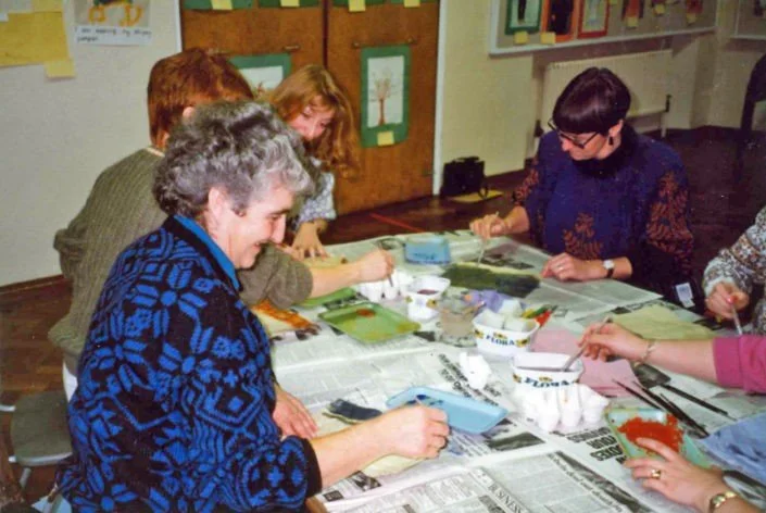 Group of women working on a craft project at a table covered with newspapers and craft supplies in a room with colorful artwork on the walls.