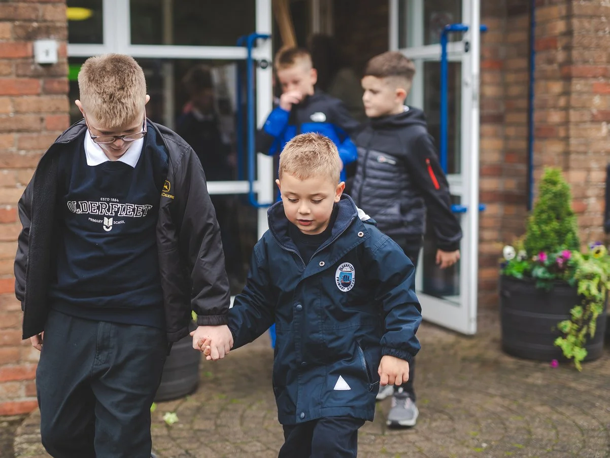 Four young boys, two holding hands, walking outside near a building with a brick wall and potted plants, wearing jackets and school uniforms.