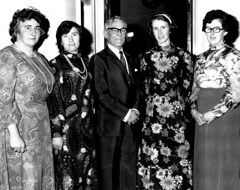 Black-and-white photo of five women and one man dressed in vintage clothing, standing indoors.