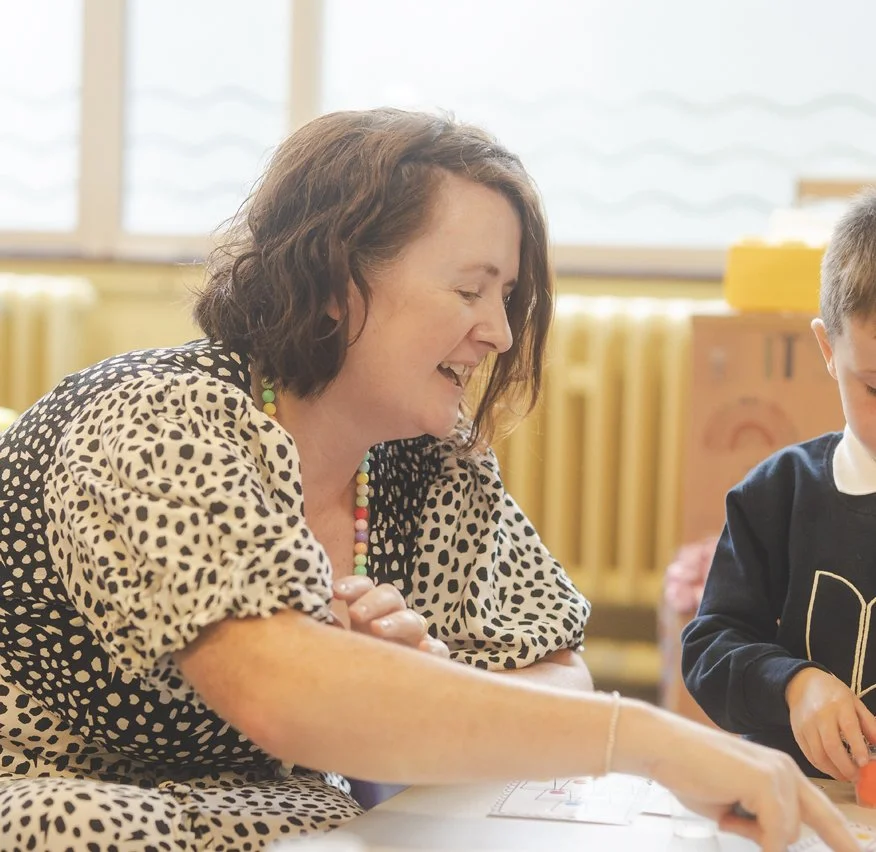 A woman smiling and engaging with a young child in a classroom setting.
