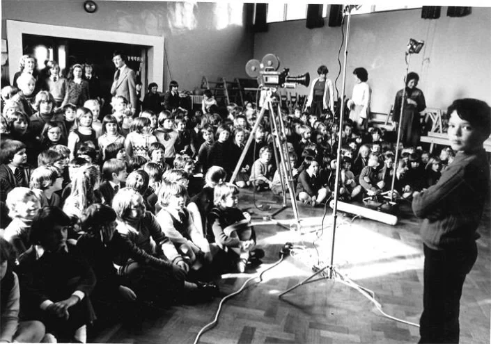 A large group of children seated on the floor in a school or community center, watching a presentation or performance. There are a few adults standing near the wall, a camera on a tripod centered in the room, and the setting looks like an indoor event or assembly.