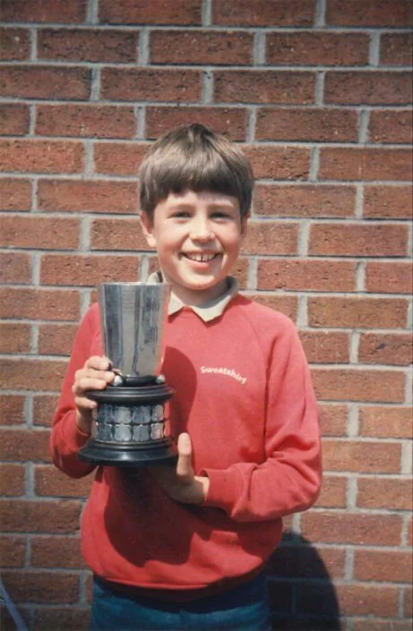 A young boy in a red sweater smiling and holding a shiny silver trophy in front of a brick wall.