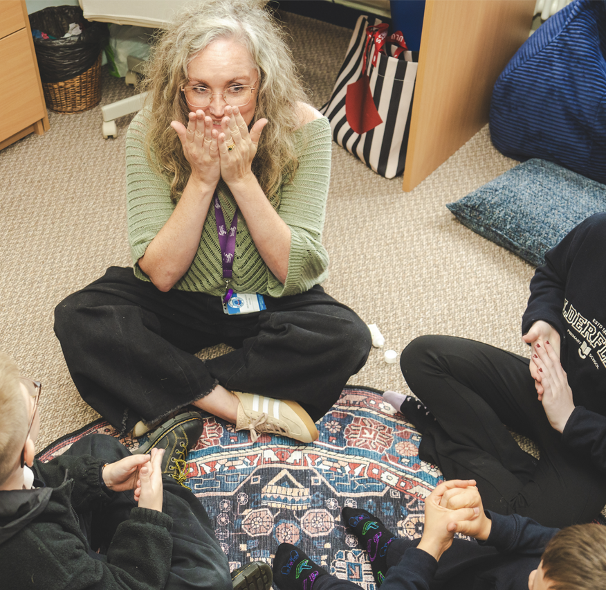 A woman with curly gray hair, glasses, and a green sweater sits cross-legged on a carpeted floor, covering her mouth with her hands, surrounded by children. The scene appears to be in a classroom or therapy setting.