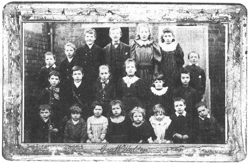 A black-and-white class photo of young children and a teacher, likely from the early 1900s, taken outside or in a school setting with a brick wall background.