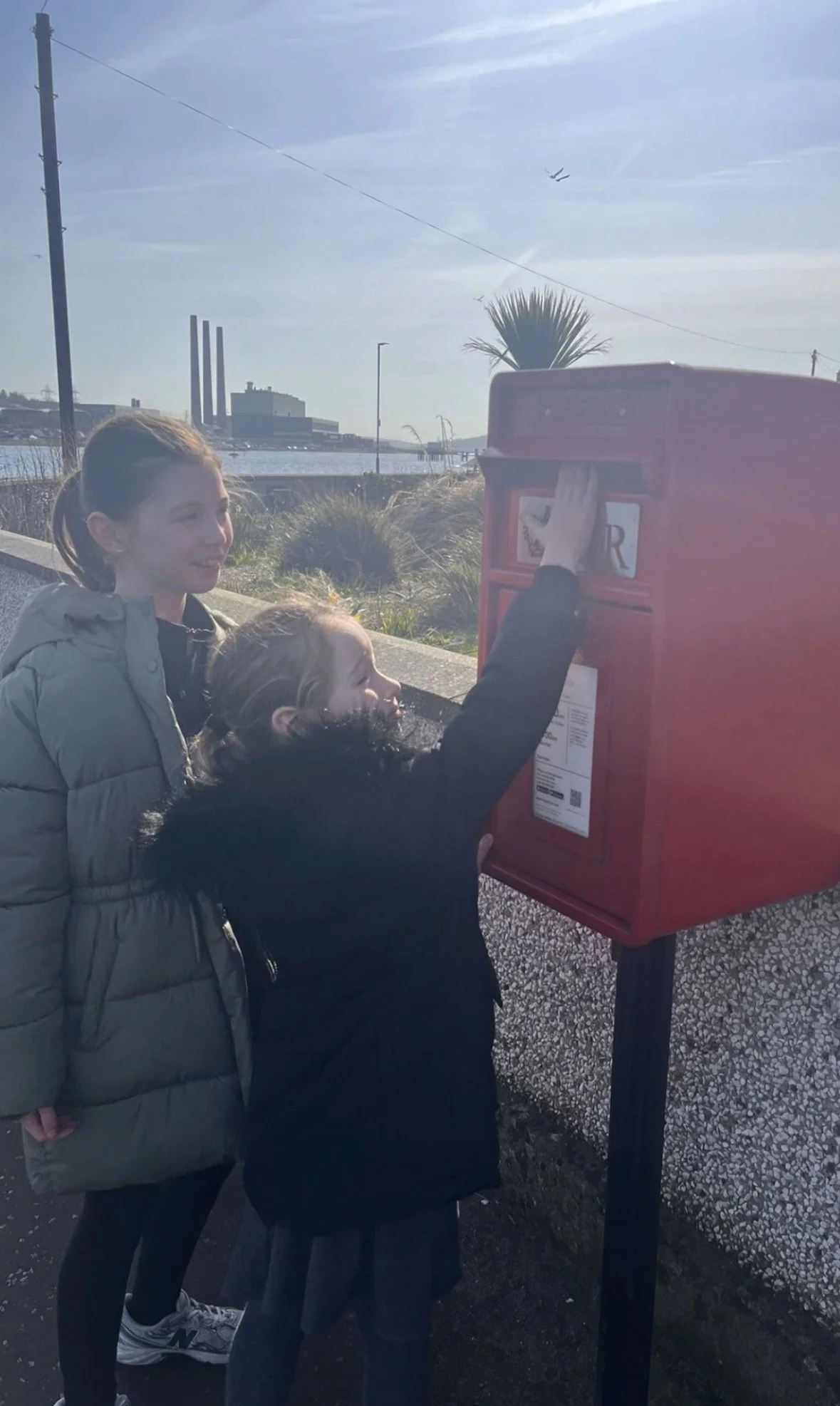 Two young girls interacting with a red mailbox near a waterfront, with industrial buildings and airplanes flying in the sky in the background.