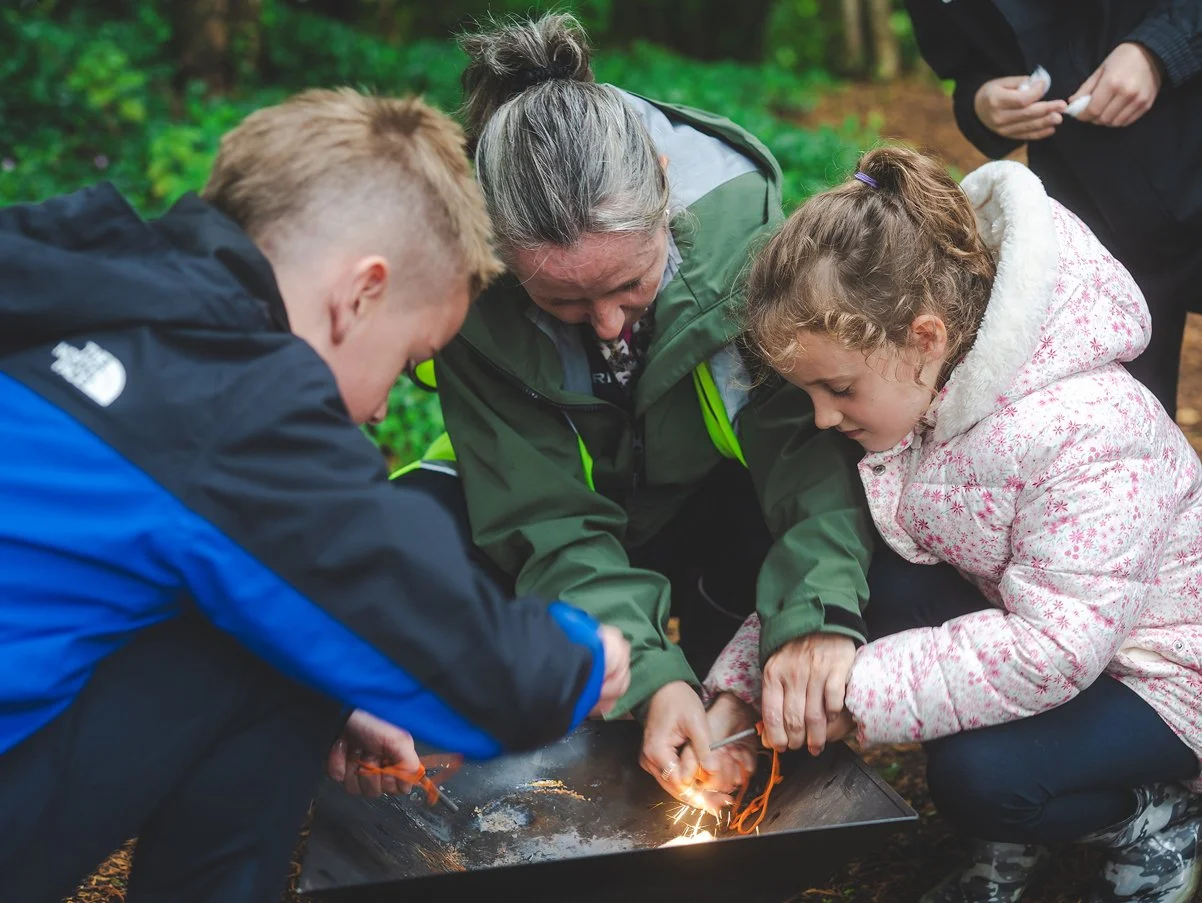Children and an adult camping outdoors, working together to start a fire with sparks flying from a metal container.
