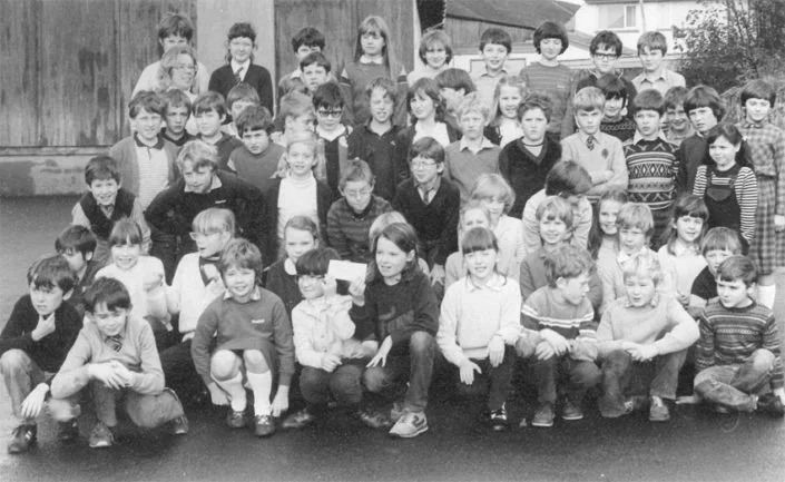 Black and white group photo of school children outdoors, arranged in multiple rows with a teacher or adult in the middle front, in a schoolyard with a fence and building in the background.