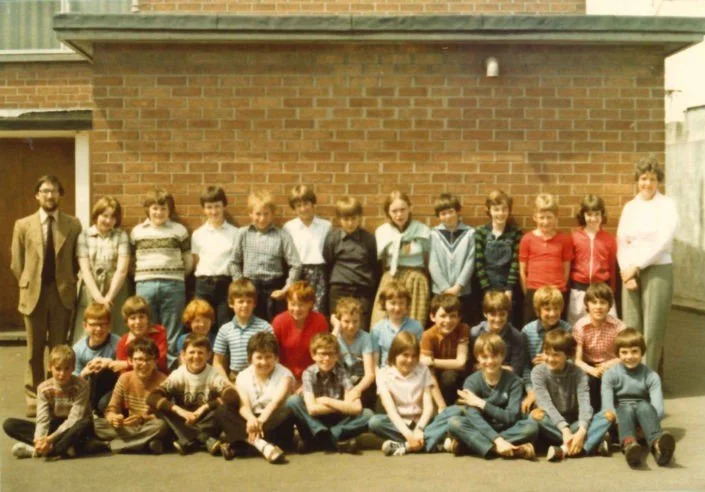 A group of children and two adults standing and sitting outside in front of a brick wall.