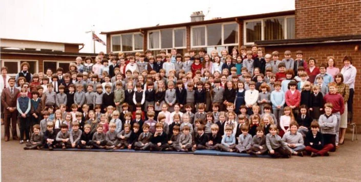 Large group of students and teachers gathered outdoors for a school photo, with a brick school building in the background.