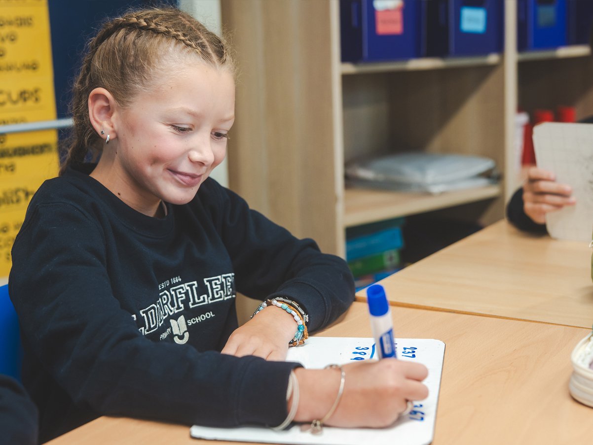 A young girl with braided hair is sitting at a desk, smiling as she writes on a whiteboard with a blue marker.