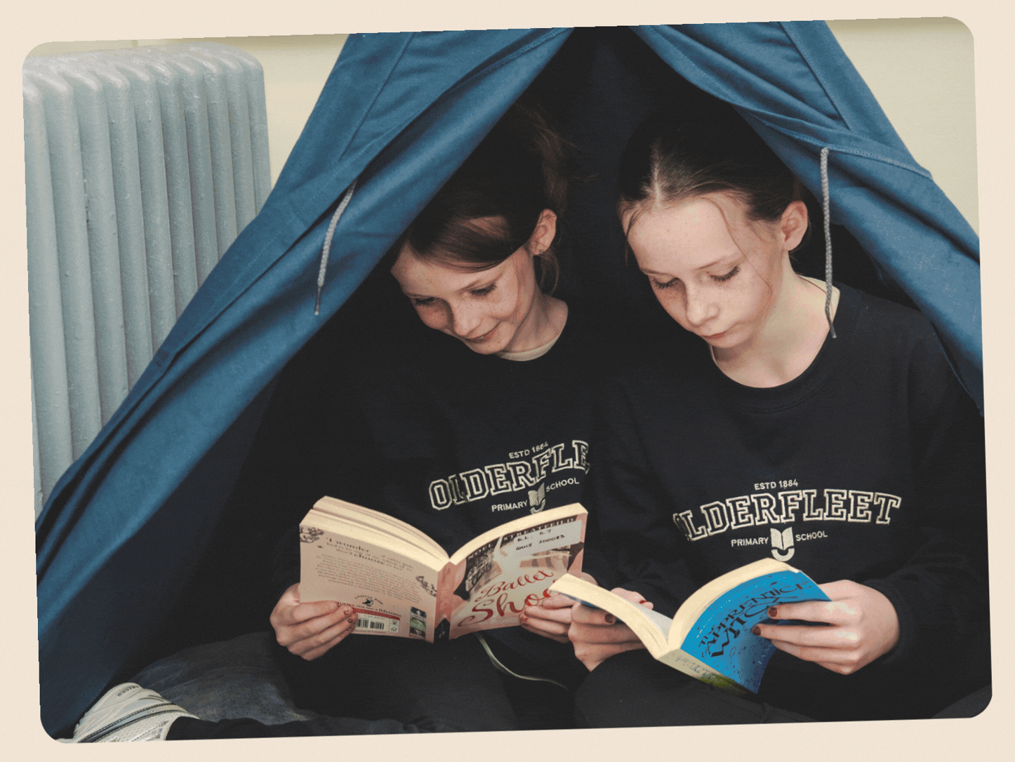 Two girls sitting under a blue tent, reading books, wearing matching black shirts with 'Olderfleet Primary School' printed on them.