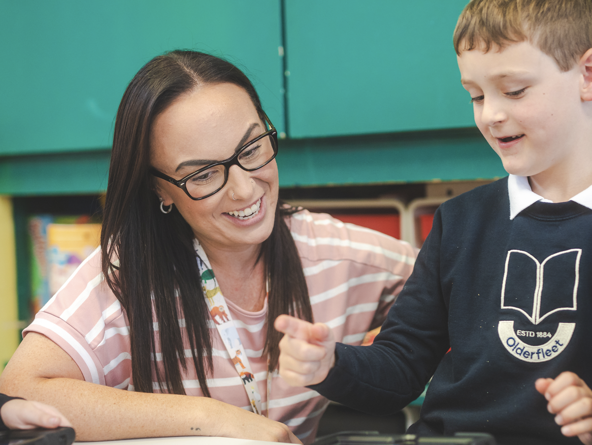 A woman with glasses and dark hair smiling and engaging with a boy wearing a navy blue school uniform with an 'Olderfleet' crest, in a classroom setting.