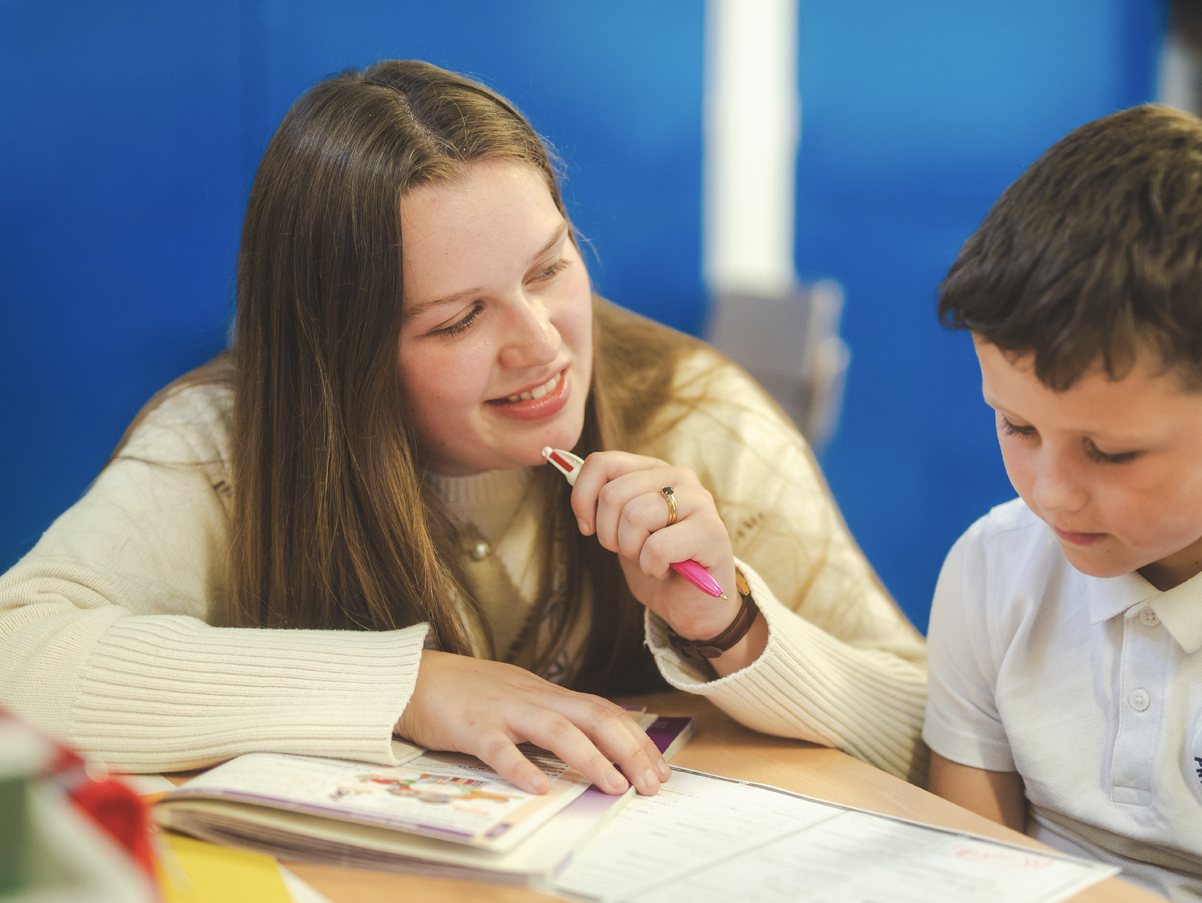 A woman with brown hair and a beige sweater helping a young boy with light brown hair and a white shirt with schoolwork at a desk.