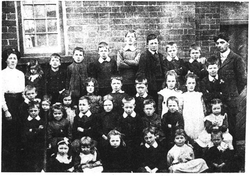 Black and white photo of a group of children and a few adults outside in front of a brick building.