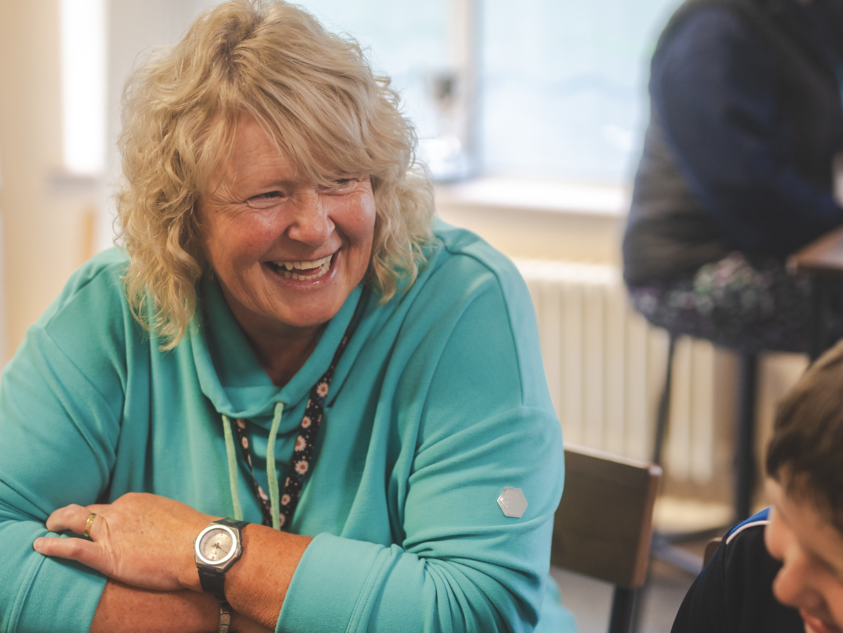 A middle-aged woman with curly blonde hair, wearing a teal hoodie, laughing in a classroom setting.