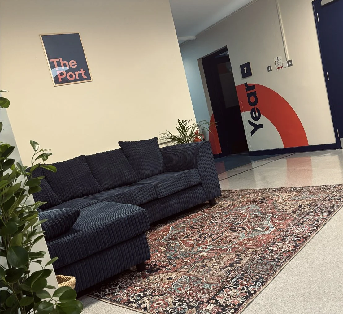 Office lounge area with a dark blue striped sofa, potted plants, a colorful patterned rug, a sign on the wall that says "The Port," and a wall mural with the words "Yecar" and "The Port."