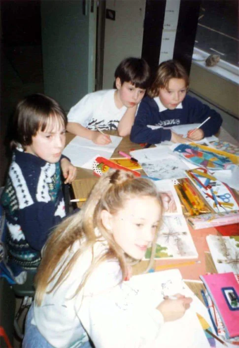 Four children sitting at a table with coloring books, crayons, and papers, engaged in drawing or coloring activities.