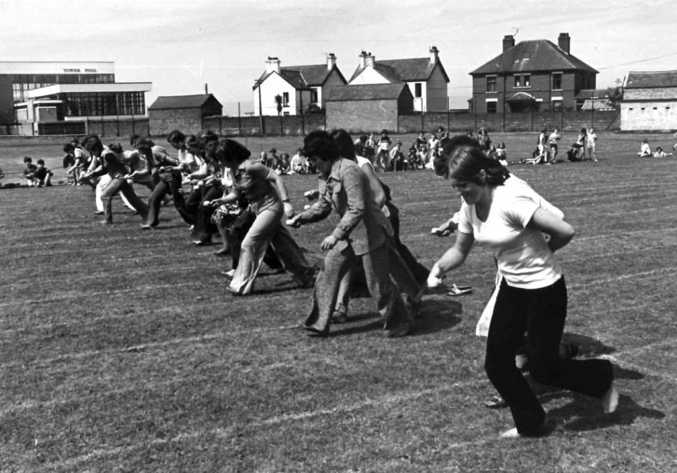 Group of people, mostly women and girls, participating in a tug-of-war game on a grassy field with houses and a building in the background.