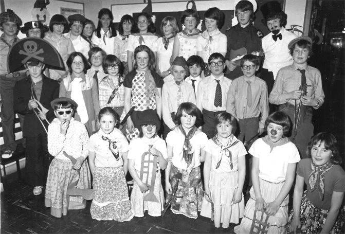 Group of children dressed in costumes for a school play or event, some wearing glasses, hats, and holding props like umbrellas and wooden chairs, standing in a classroom or auditorium.