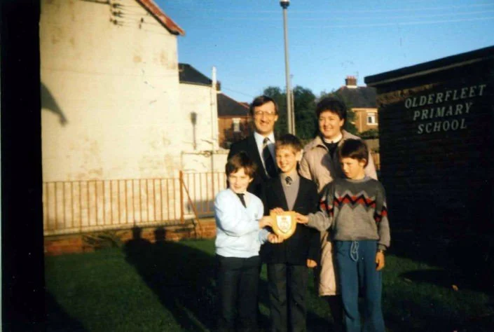A group of five children and one adult standing outside a school building, holding a shield-shaped plaque, in front of a sign that reads 'Olderfleet Primary School.'