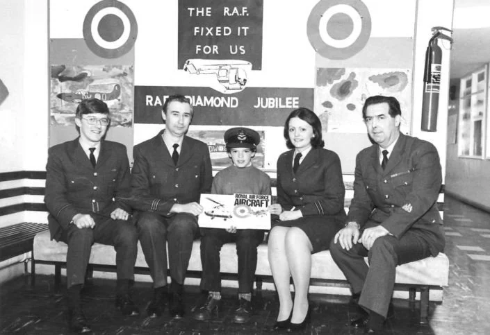 A group of four uniformed RAF personnel and a young child in the center holding a model airplane, seated on a bench in a decorated room with RAF insignia and a banner celebrating the Royal Air Force Diamond Jubilee.