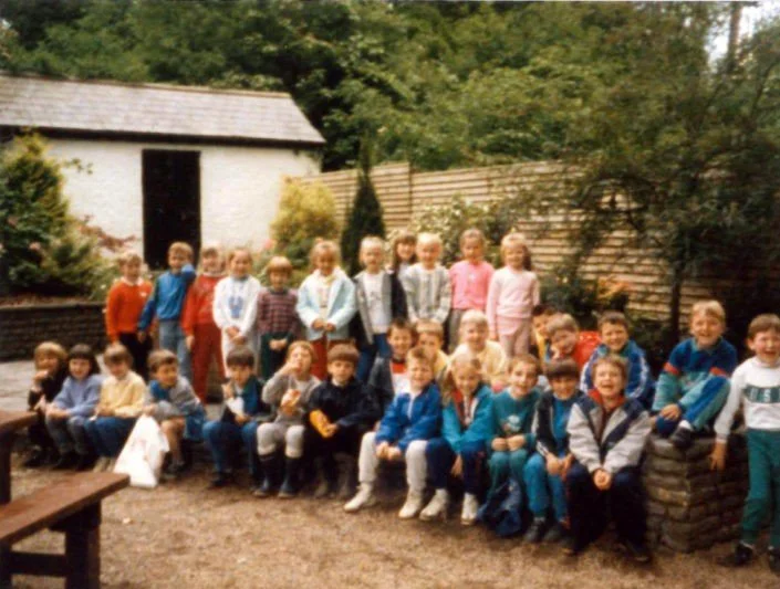 Group of children outdoors in a garden or backyard poses for a photo, some sitting on a bench and some standing, with trees and a wooden shed in the background.