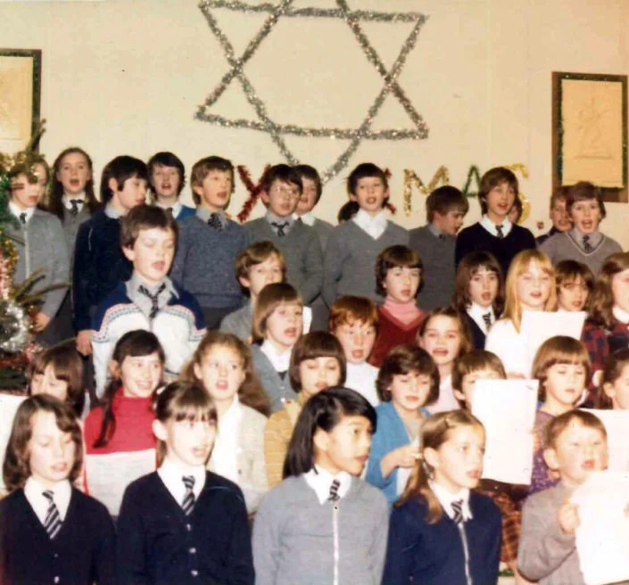 Group of children singing or performing on stage with a Star of David made of tinsel in the background, decorated for Hanukkah celebration.