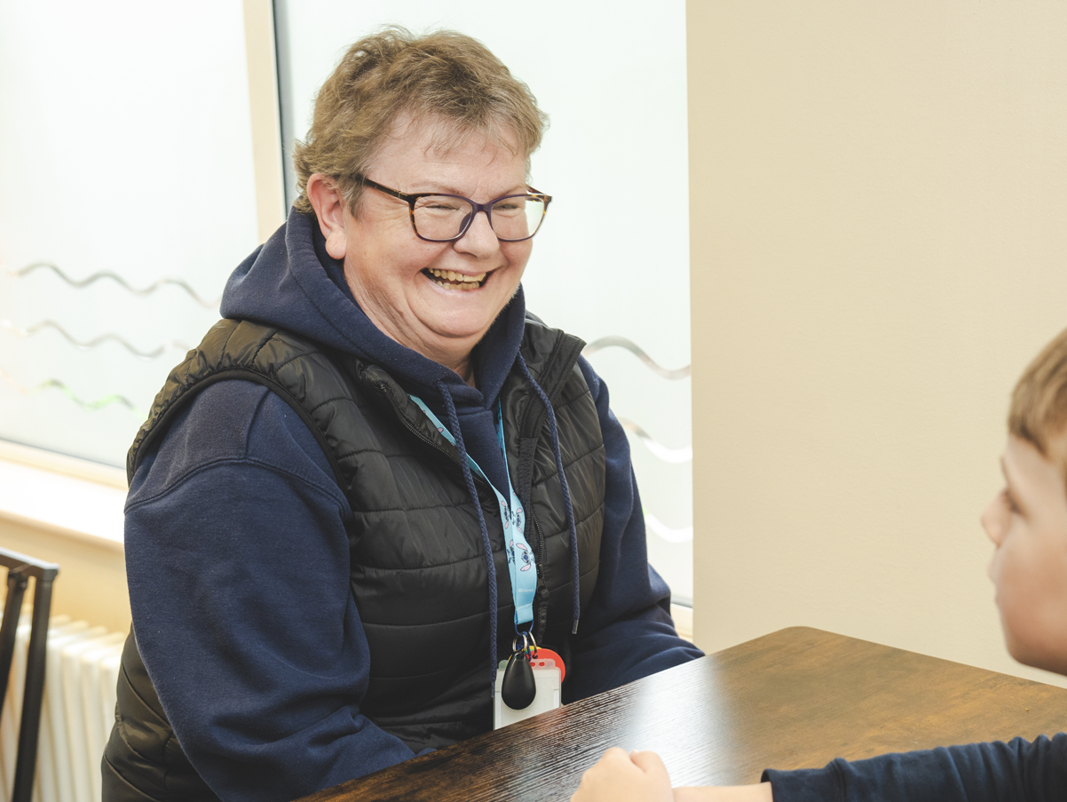 A smiling woman with glasses and a vest sitting at a table talking to a young boy.