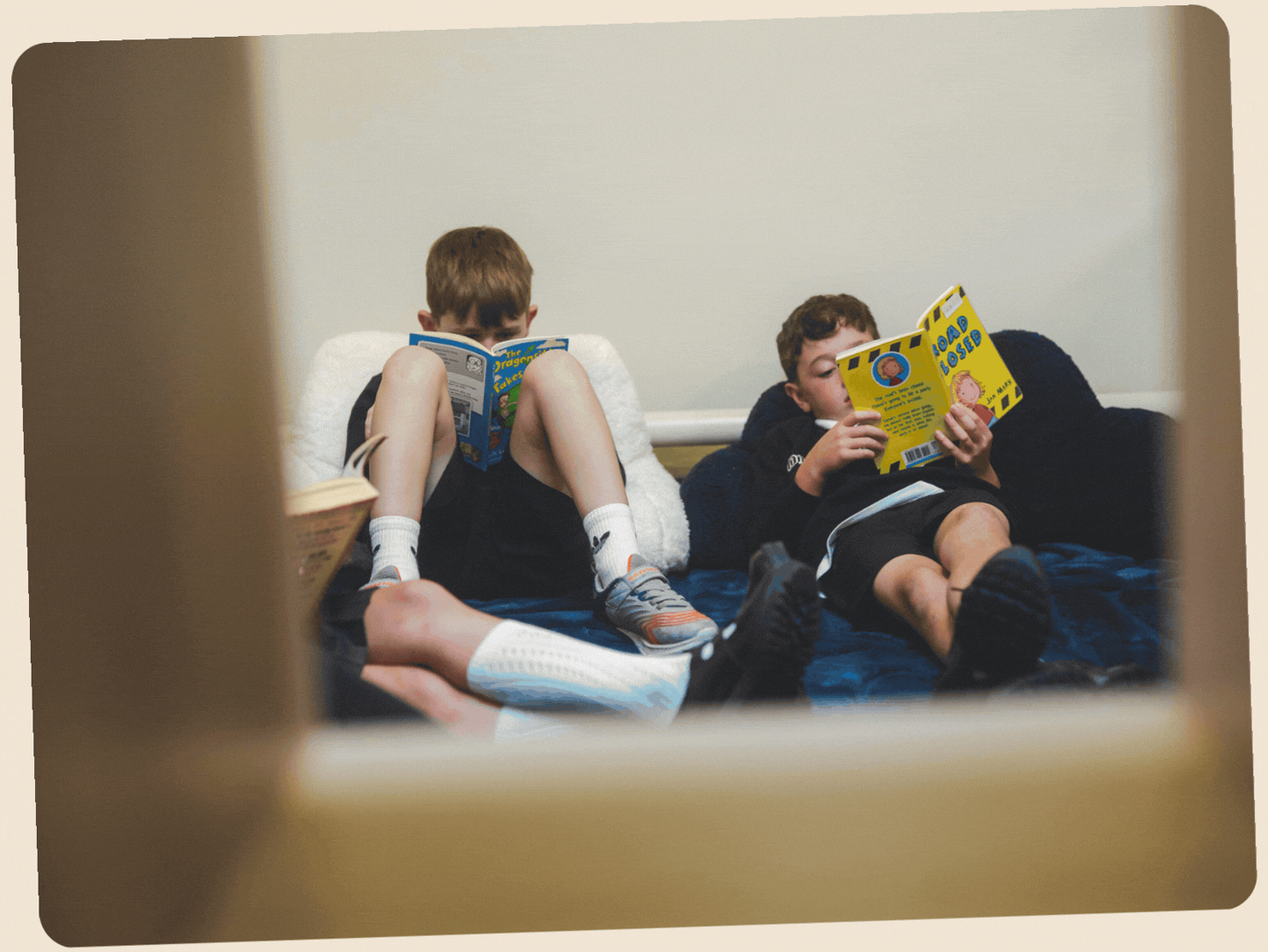 Two boys are lying on a bed, each reading a book. The boy on the left has short brown hair and is wearing a black shirt and shorts, with striped socks and sneakers. The boy on the right has curly brown hair and is wearing a black shirt and black shorts. They are both relaxed, reading their respective books.