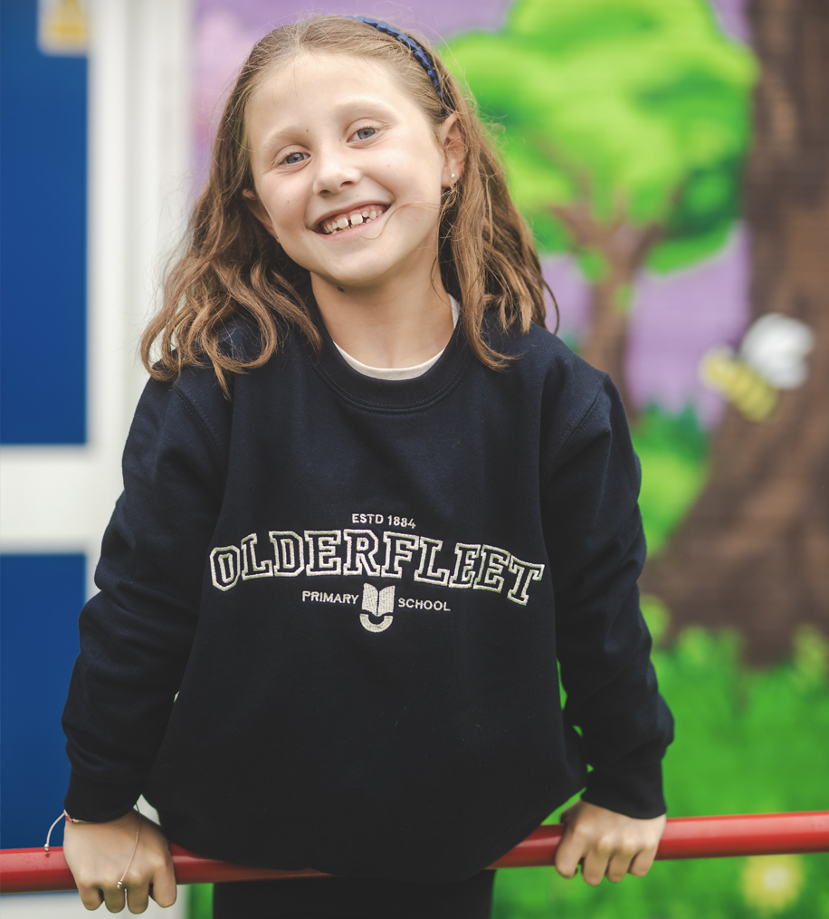 A smiling young girl with brown curly hair and a blue headband, wearing a black Oldersfleet Primary School sweatshirt, holding her hands on a red railing with a colorful mural and tree in the background.